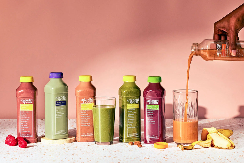Assorted Splendid Spoon Smoothie bottles with colorful lids on a pink background, with a hand pouring juice into a glass.