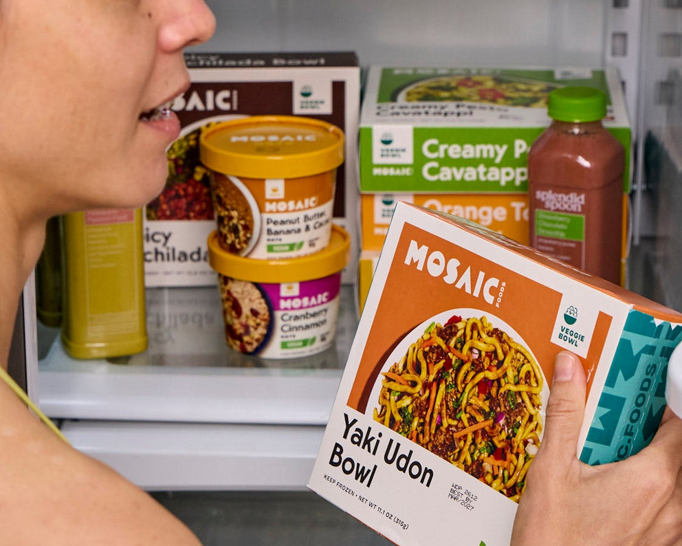 Woman holding a Mosaic Foods package in front of an open refrigerator filled with various food items.