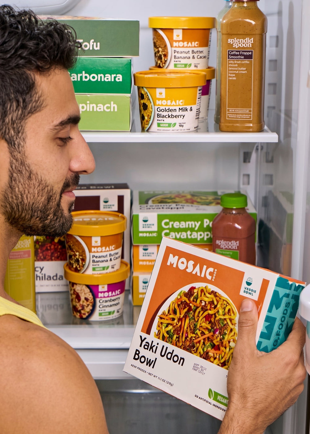 Man looking at a Mosaic Foods Yaki Udon Bowl package in front of a refrigerator filled with various food items.