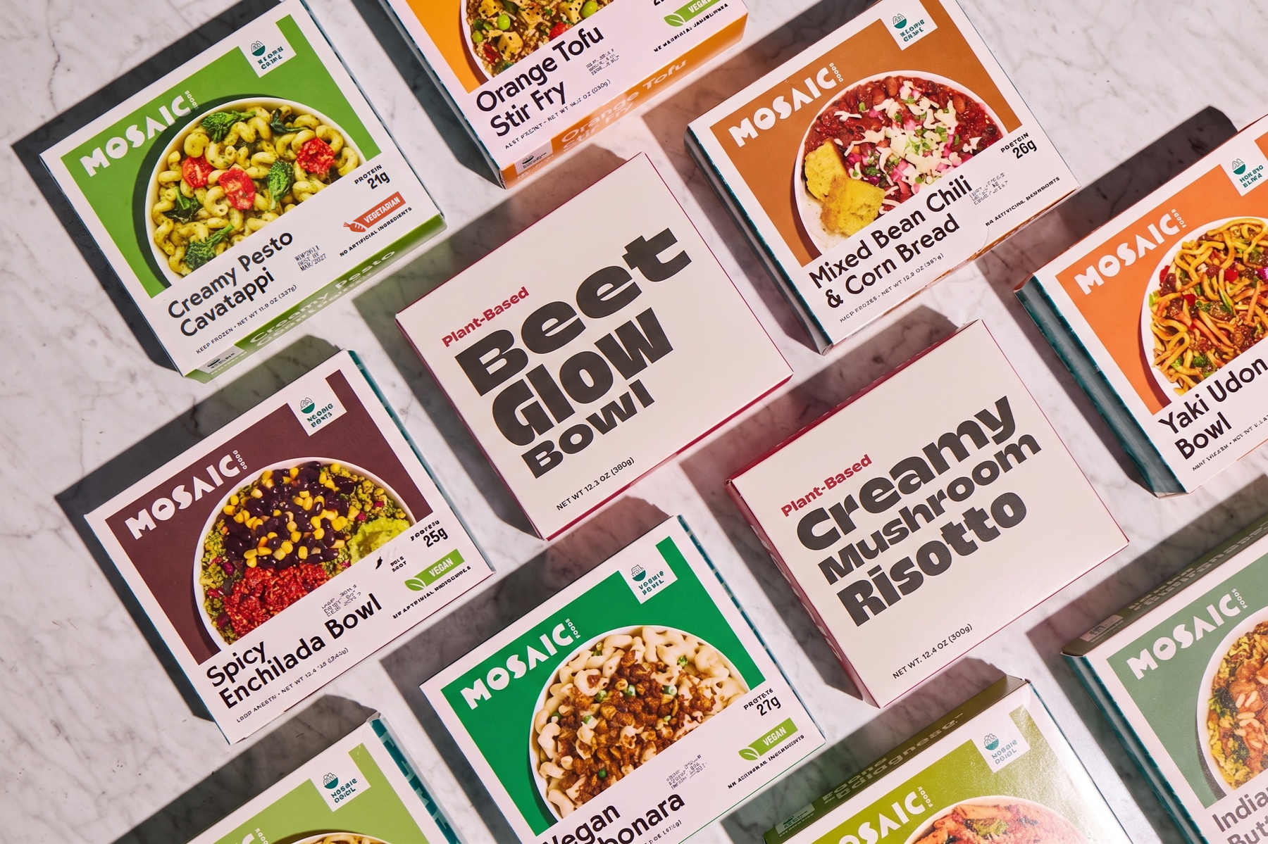 Several Mosaici Plant-Based meal kit boxes arranged on a light marble surface. Visible flavors include Beet Glow Bowl, Creamy Mushroom Risotto, Spicy Enchilada Bowl, Orange Tau Sli Fry, Creamy Keto Cassapaya, Mixed Bean Chili & Corn Bread, and Yaki Udon Bowl.